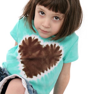 Peut inclure: Une jeune fille portant un t-shirt turquoise tie-dye avec un motif de cœur brun. Elle porte un short en jean avec une bordure en dentelle blanche.