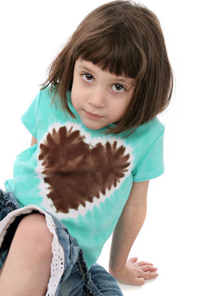 May include: A young girl wearing a turquoise tie-dye t-shirt with a brown heart design. She is wearing denim shorts with white lace trim.