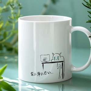 Könnte beinhalten: Eine weiße Keramiktasse mit einer Schwarzweißillustration einer Katze an einem Schreibtisch mit einem Computer. Die Katze hat blaue Augen und sieht traurig aus. Der Text auf der Tasse lautet "家に帰りたい...", was übersetzt "Ich möchte nach Hause..." bedeutet.