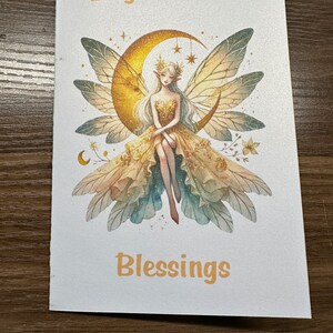 May include: A white card with a gold glitter background featuring a watercolor illustration of a fairy sitting on a crescent moon. The fairy has long blonde hair, a gold crown, and a gold dress. The text "Brightest Litha Blessings" is written in gold lettering.
