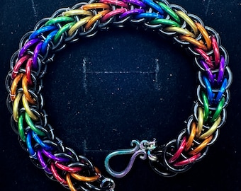 May include: A rainbow colored chainmail bracelet with a silver clasp. The bracelet is made of small metal rings that are linked together in a pattern.