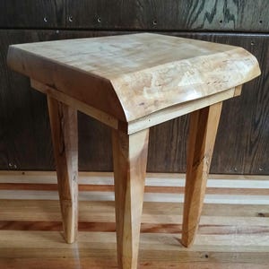 May include: A small, handcrafted wooden side table with a light-colored, natural wood finish. The table has a square top with rounded edges and four angled legs. The wood grain is visible, adding to its rustic charm. The table is approximately 18 inches tall.