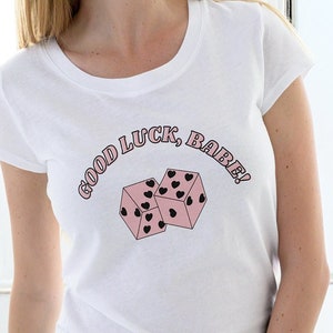 Puede incluir: Camiseta blanca con cuello redondo y mangas cortas. La camiseta presenta el texto "GOOD LUCK, BABE!" en rosa, en forma de arco, sobre dos dados rosas con puntos de corazón negros. Los dados están ligeramente inclinados.
