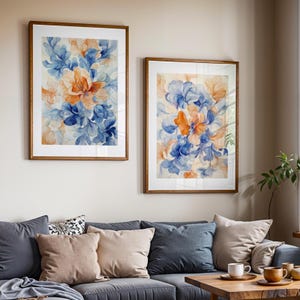 May include: Two framed floral art prints. Each print features a watercolor design with blue, orange, and cream-colored flowers and leaves. The frames are a warm brown color. The prints are hanging above a gray sofa with pillows.