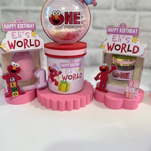 May include: A pink and red Sesame Street-themed birthday decoration set. The set includes a central container with an Elmo graphic, a clear globe with the word "ONE", and additional decorative elements featuring Elmo and Abby Cadabby.