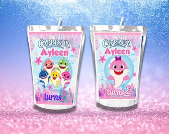May include: Two Capri Sun juice pouches with straws. One features a group of cartoon sharks, the other a single pink shark. Both pouches have the name "Ayleen" and the words "turns 2". Pink and blue background.