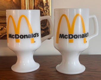May include: Two white ceramic mugs with a yellow and black McDonald's logo. The mugs have a pedestal base.
