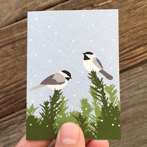 May include: A blue and white card with a snowy background featuring two black-capped chickadees perched on green pine branches.