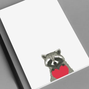 May include: A white notepad with a raccoon illustration holding a red heart. A black pencil rests on the gray surface next to the notepad. The raccoon is gray and white with a black mask.