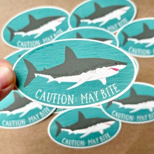 May include: A set of eight oval-shaped stickers with a turquoise background and a white great white shark illustration. The stickers feature the text "CAUTION: MAY BITE" in white lettering.