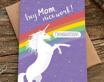 Puede incluir: Tarjeta de felicitación con fondo morado, arcoíris y unicornio blanco. El texto de la tarjeta dice "hey Mom, nice work!" y "I'M MAGNIFICENT." La tarjeta está sobre una superficie de madera con un sobre marrón y lápices.