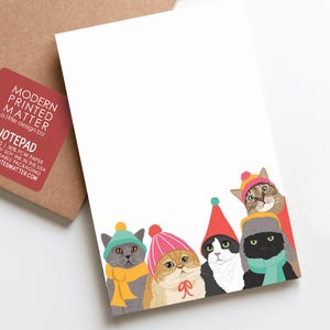 May include: A white notepad featuring a colorful illustration of five cats wearing winter hats and scarves at the bottom. The cats include a grey cat, a ginger cat, a black and white cat, and a black cat. A red label with white text is visible.