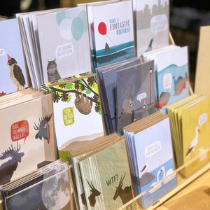 以下が含まれることがあります： ユーモラスなイラストとテキストが描かれたグリーティングカードのディスプレイ。カードには、ナマケモノ、ヘラジカ、ユニコーン、鳥など、さまざまな動物が描かれています。カードのテキストには、「ゆっくりしろよ、くそったれ」や「枕に毛玉を置いてきたよ」や「月まで行って帰ってこい」などのフレーズが書かれています。