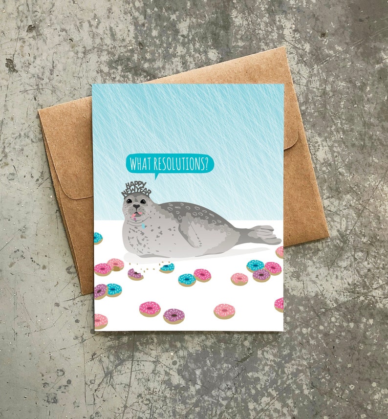 May include: A greeting card featuring a cartoon seal wearing a "Happy New Year" crown and surrounded by pink and blue doughnuts. The seal has a speech bubble that says "What Resolutions?"