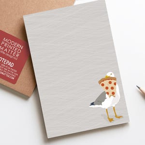 Könnte beinhalten: Notizblock mit hellgrauem Hintergrund mit einer Cartoon-Möwe, die ein Pizzastück trägt. Ein roter Aufkleber mit weißem Text "MODERN PRINTED MATTER" befindet sich auf einem braunen Kartonträger. Ein Bleistift befindet sich auf der rechten Seite.