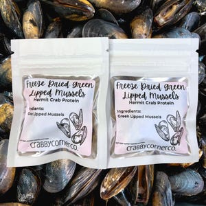 May include: Two small, clear pouches of freeze-dried green lipped mussels, a hermit crab protein. Each pouch has a pink label with text and a mussel illustration. The background is filled with mussel shells.