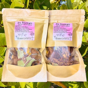 May include: Two beige pouches labeled "Sea Scavenger" for hermit crab foraging. Each pouch contains a mix of dried leaves and botanicals. The ingredients listed include sea grape leaves, sunflowers, and seaweed. The brand name "Crabbycornerco." is visible.
