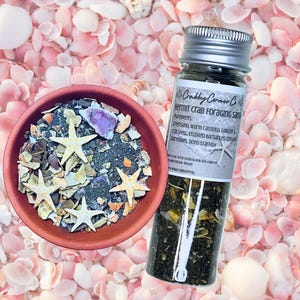 May include: A small terracotta bowl filled with dark foraging sand, crushed shells, and four small white starfish, alongside a clear glass vial with a silver lid containing the same mixture, labeled Crabby Corner Co. Hermit Crab Foraging Sand.