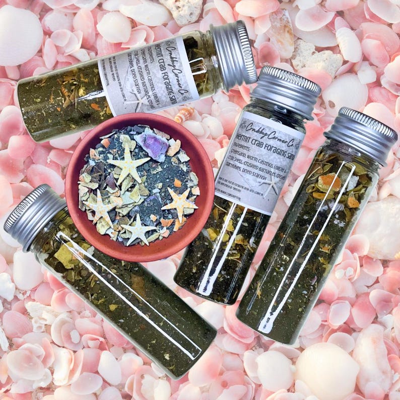 May include: Three clear glass vials with silver screw-top lids containing a dark green granular mixture labeled Crabby Corner Co. Hermit Crab Foraging Sand, alongside a small terracotta bowl filled with a mix of sand, shells, and starfish.