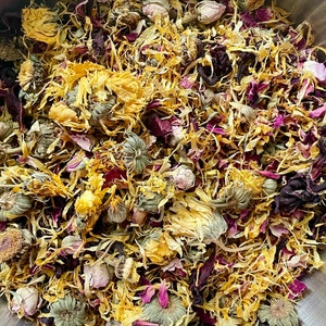 May include: A bowl filled with a mixture of dried flowers and herbs, including yellow calendula petals, pink rose petals, and green herbs.