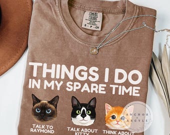 May include: Espresso-colored t-shirt with the text "THINGS I DO IN MY SPARE TIME" and three cat illustrations. The shirt also features the phrases "TALK TO RAYMOND", "TALK ABOUT KITTY", and "THINK ABOUT BOB". A silver necklace is draped across the shirt.