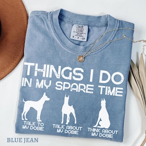 May include: A blue jean t-shirt with white text that reads "Things I do in my spare time". The shirt features three silhouettes of Doberman Pinschers with the text "Talk to my Dober", "Talk about my Dober", and "Think about my Dober" underneath each silhouette.