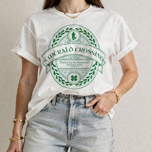 Può includere: T-shirt bianca con maniche arrotolate, con un design circolare verde con la scritta "EMERALD CROSSING". Il design include un trifoglio e la citazione "The Lord is my shepherd; I shall not want. Psalm 23:1".