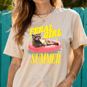 May include: Beige t-shirt featuring a raccoon wearing sunglasses and floating on a pink inflatable ring. The text "FERAL GIRL SUMMER" is printed in yellow above and below the raccoon illustration. The shirt has short sleeves.