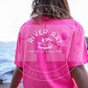Puede incluir: Camiseta rosa brillante con un gráfico blanco que dice "RIVER RAT" sobre una ilustración de una rata de dibujos animados en un flotador. El texto "FUELED BY SUN & FUN" y "est. 2020" también están impresos en la camiseta. Las palabras "play outdoor" están impresas en un círculo.