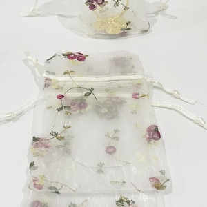 Puede incluir: Bolsa de tela blanca transparente con un patrón floral. La bolsa tiene un cierre de cordón y está hecha de un material delicado y translúcido.