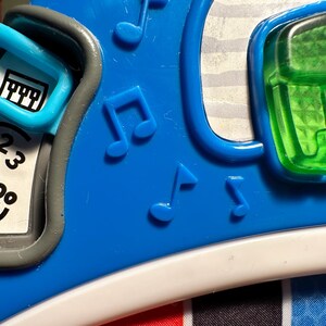 May include: A blue plastic toy featuring a sliding switch with a piano icon, a white panel with the numbers 1, 2, 3 and a paw print icon, embossed musical note symbols, and a translucent green rectangular button with a textured grid pattern.