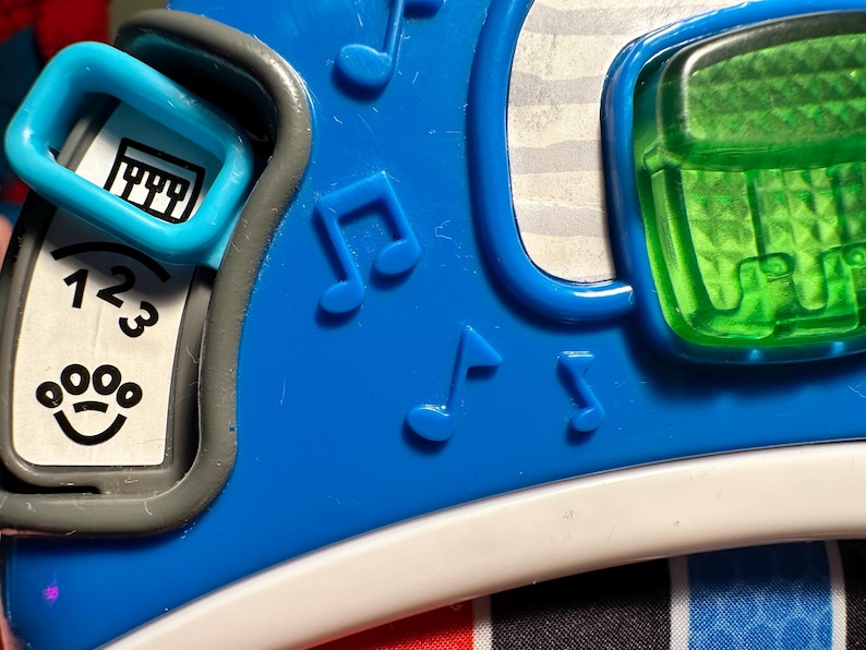 May include: A blue plastic toy featuring a sliding switch with a piano icon, a white panel with the numbers 1, 2, 3 and a paw print icon, embossed musical note symbols, and a translucent green rectangular button with a textured grid pattern.