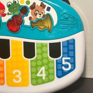 May include: A turquoise and white musical toy piano featuring a top panel with illustrations of a red bird, a green frog, and a brown dog. The piano keys are labeled with the numbers 3, 4, and 5, and include translucent red violin and yellow trumpet icons.