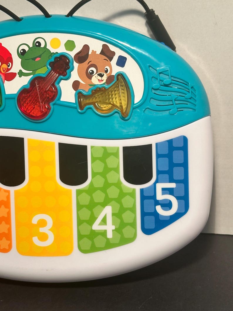 May include: A turquoise and white musical toy piano featuring a top panel with illustrations of a red bird, a green frog, and a brown dog. The piano keys are labeled with the numbers 3, 4, and 5, and include translucent red violin and yellow trumpet icons.