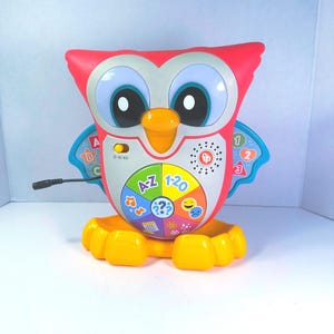 May include: A Fisher-Price electronic owl toy with a red body, yellow feet, and a central circular interface featuring A-Z, 1-20, and various icons. The owl has large white eyes, an orange beak, and blue wings with letters and numbers printed on them.