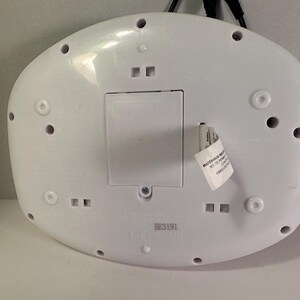May include: A white, oval-shaped plastic electronic device base featuring a central rectangular battery compartment cover, multiple circular screw holes, and a small white product tag with printed text attached to the side of the casing.