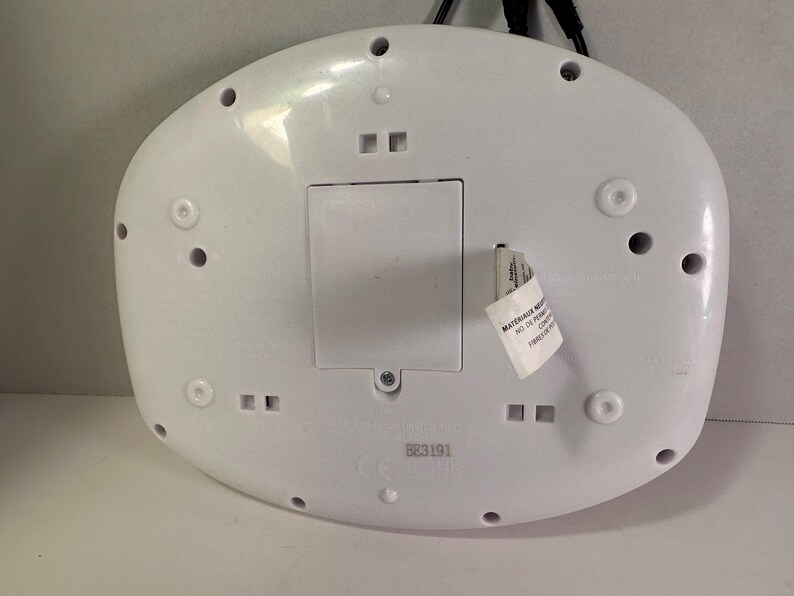 May include: A white, oval-shaped plastic electronic device base featuring a central rectangular battery compartment cover, multiple circular screw holes, and a small white product tag with printed text attached to the side of the casing.