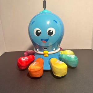 May include: A blue plastic octopus toy with a smiling face, a small black antenna on top, and a white collar. The octopus has eight soft, colorful fabric tentacles, each featuring a printed white number, including 8, 7, 4, and 3.