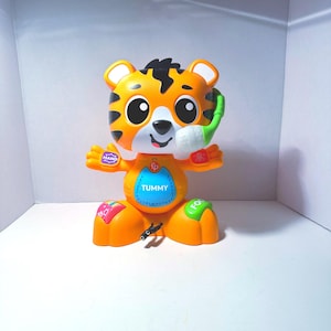 May include: An orange and black plastic tiger toy with large eyes, a white muzzle, and a green microphone attachment. The toy features a blue center panel labeled TUMMY with alphabet letters, and buttons on the hands and feet labeled HAND and FOOT.