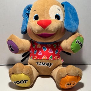 May include: A plush, interactive puppy toy with a tan body, blue ears, and a red nose. The puppy wears a red shirt with colorful letters and the word "TUMMY". The paws and hands have the words "FOOT" and "HAND" printed on them. The puppy has a white spot on its face.