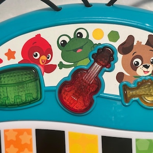 May include: A teal plastic musical toy console featuring a white panel with illustrations of a red bird, a green frog, and a brown dog. Three translucent buttons shaped like a green drum, a red guitar, and a yellow trumpet are integrated.