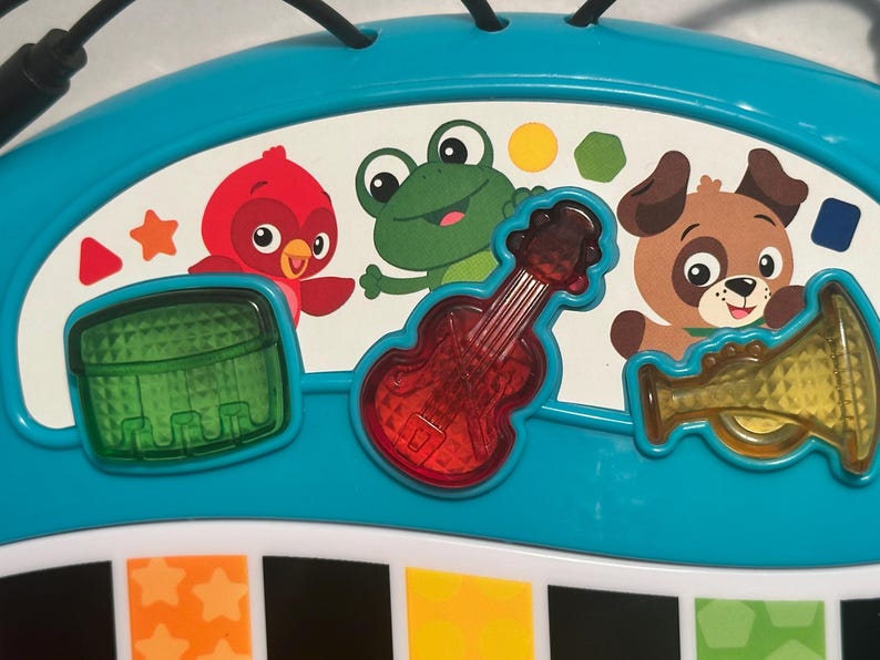 May include: A teal plastic musical toy console featuring a white panel with illustrations of a red bird, a green frog, and a brown dog. Three translucent buttons shaped like a green drum, a red guitar, and a yellow trumpet are integrated.