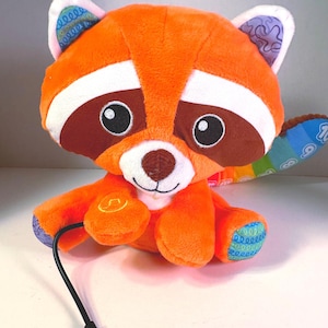 May include: Orange plush raccoon toy with a rainbow tail and a black cord. The raccoon has white fur around its eyes and a brown nose. The toy has a button on its chest.