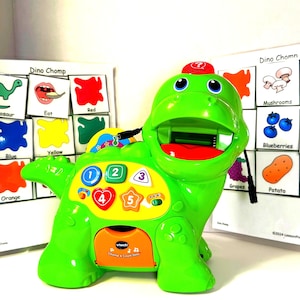 May include: A green dinosaur-shaped toy with a red mouth and a red hat. The toy has buttons with numbers and shapes. Two educational boards with pictures of food and colors are behind the toy. The text "Dino Chomp" is visible.