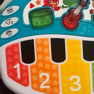 May include: A teal and white plastic musical toy with a piano keyboard featuring red, orange, and yellow keys labeled with the numbers 1, 2, and 3. The top section displays a red bird, a green frog, a green drum, and a red violin icon.