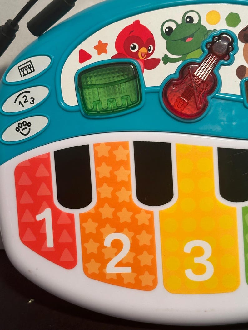 May include: A teal and white plastic musical toy with a piano keyboard featuring red, orange, and yellow keys labeled with the numbers 1, 2, and 3. The top section displays a red bird, a green frog, a green drum, and a red violin icon.