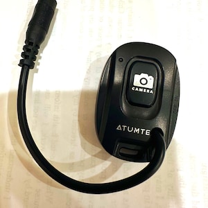 May include: A black camera remote control with a button labeled "CAMERA" and the brand name "ATUMTE". The remote has an attached black cable and connector. The on/off switch is visible on the side.