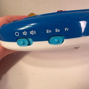 May include: A blue and white electronic device featuring two light blue sliding switches. The left switch adjusts volume settings indicated by a circle and speaker icons. The right switch selects languages labeled as En, Es, and Fr in white text.