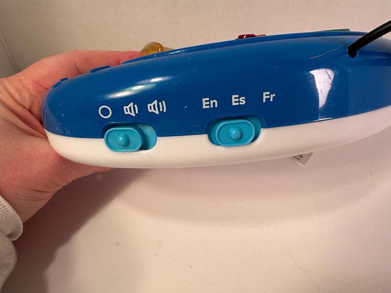 May include: A blue and white electronic device featuring two light blue sliding switches. The left switch adjusts volume settings indicated by a circle and speaker icons. The right switch selects languages labeled as En, Es, and Fr in white text.