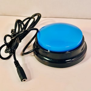 May include: A circular blue push button switch with a glossy finish, mounted on a black plastic base. A black electrical cord extends from the side of the base, terminating in a standard cylindrical audio-style jack connector for device input.